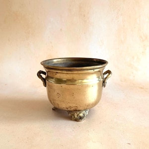 May include: A polished brass pot featuring a wide circular opening, two small side handles, and three decorative feet. The metal surface displays a warm golden hue with subtle signs of age and wear, including minor surface imperfections.