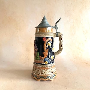 May include: A vintage ceramic beer stein with a hinged metal lid. The stein features a dark blue body with floral and figural relief designs, a decorative handle, and the word "MIR" on the side. The base has an oak leaf and acorn design.