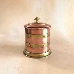 May include: A cylindrical copper canister featuring two horizontal brass bands secured with small rivets. The container includes a matching copper lid topped with a small brass knob and a wide brass base, creating a metallic storage vessel.