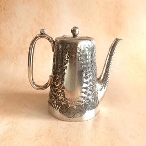 May include: A silver-toned metal teapot featuring an engraved botanical pattern of ferns and leaves around the body. The teapot has a tall, cylindrical shape, a curved handle with a small knob at the top, a lid with a spherical finial, and a spout.
