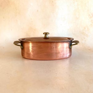 May include: An oval copper casserole dish with a lid and brass handles. The dish has a shiny surface and a small brass knob on the lid. The handles are curved. A classic kitchenware item. The dish is approximately 25 cm in length.