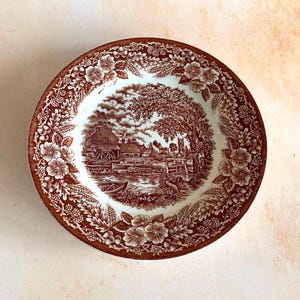 May include: A round ceramic plate with a brown and white landscape scene in the centre. The rim has a floral and foliage pattern. The plate's design has a vintage aesthetic, suitable for serving or display, reminiscent of classic tableware.
