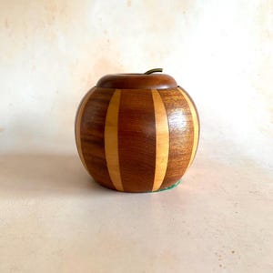 Könnte beinhalten: Eine runde Holzbox mit Deckel, mit abwechselnden vertikalen Streifen aus dunklem und hellem Holz. Der Deckel hat einen kleinen, gebogenen Metallgriff. Die Box ist so gestaltet, dass sie einem Apfel ähnelt.