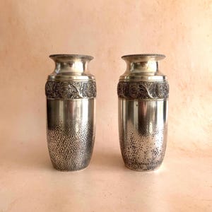 Könnte beinhalten: Zwei silberne Metallvasen. Jede Vase hat einen strukturierten, gehämmerten unteren Abschnitt und ein dekoratives Band mit floralem Relief. Ober- und Unterseite haben eine glatte, polierte Oberfläche. Die Vasen sind zylindrisch und klassisch gestaltet.