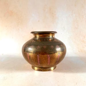 May include: A round brass pot with a wide opening and a short neck. The exterior features intricate etched patterns, including vertical panels and diamond shapes, with alternating sections of polished brass and copper-toned metal accents.