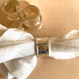 May include: Silver napkin rings with an ornate, scrolling design. One ring holds a white linen napkin, while others are stacked nearby. The rings have a polished finish. A classic table setting accessory.