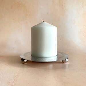 May include: A white pillar candle on a silver candle holder with a decorative edge and three small ball feet. The candle is unlit and has a black wick.