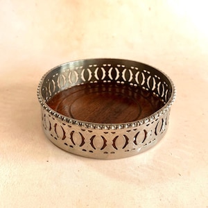 May include: A round coaster with a silver metal filigree border and a dark wood centre. The coaster has an openwork design around the edge and a beaded trim. This decorative coaster is suitable for a variety of home decor styles.