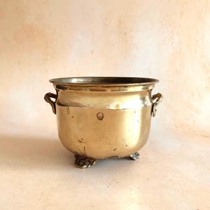 May include: A polished brass pot with a rounded body, a wide circular opening, and two small side handles. The vessel is supported by three ornate, decorative feet and features a smooth, metallic gold-toned surface with minor surface markings.