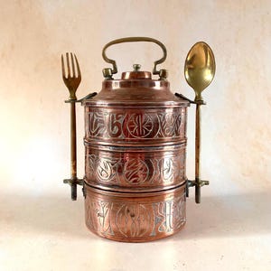 May include: A three-tiered copper tiffin carrier featuring intricate etched patterns on each cylindrical section. A brass handle sits on top, while a brass fork and a brass spoon are attached to the sides of the container with metal hardware.