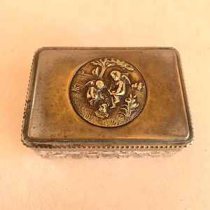 Könnte beinhalten: Eine rechteckige Schmuckschatulle mit einem klaren, facettierten Glasboden und einem angelaufenen Messingdeckel. Der Deckel zeigt ein zentrales, kreisförmiges Medaillon mit zwei geflügelten Putten in einer Landschaft mit Baum und Blumen.