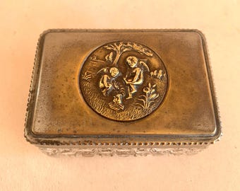 May include: A rectangular trinket box with a clear, faceted glass base and a tarnished brass lid. The lid features a central circular medallion depicting two winged cherubs in a landscape setting with a tree, grass, and small floral elements.