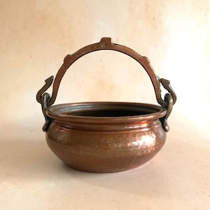 May include: A round, hammered copper bowl featuring a wide, arched metal handle attached to two decorative, curved side mounts. The bowl has a polished, metallic finish with visible indentations across its surface and a smooth, rounded rim.