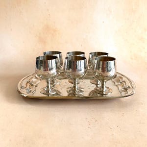 May include: A set of six silver-coloured cordial glasses with etched floral designs, presented on a matching oval tray. The glasses have a classic stemmed design, reflecting light. The tray has a decorative edge.