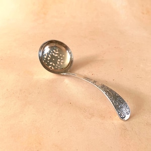 May include: A silver-toned metal slotted spoon featuring a circular bowl with a decorative pierced floral pattern. The curved handle is adorned with an intricate embossed botanical design that extends along its entire length.