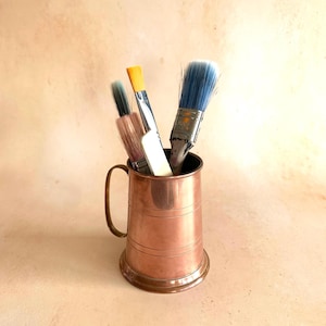 May include: A copper-coloured metal mug with a handle, containing paintbrushes. The brushes have blue, yellow, and pink bristles. The mug features horizontal lines around its body. The mug is likely used for art supplies and sits on a light-coloured surface.