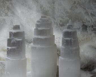 May include: Three white selenite crystal towers of varying sizes, arranged on a white textured surface.