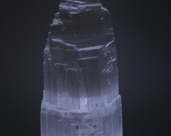 May include: A white selenite crystal tower with a layered, stacked design. The crystal is illuminated from below, creating a soft glow.