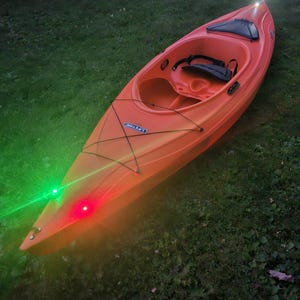May include: An orange Lifetime kayak with a black seat and storage area. The kayak has red and green lights on the front and a white light on the back. The kayak is sitting on green grass.