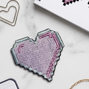 May include: A pixelated heart-shaped patch in shades of lavender, pink, and teal, with a black border. The patch is embroidered and has a textured appearance. The design is reminiscent of retro video game graphics.