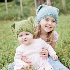 Può includere: Due ragazze con cappelli di lana, uno verde e uno blu, sono sedute in un campo erboso. La ragazza con il cappello verde tiene la ragazza con il cappello blu.