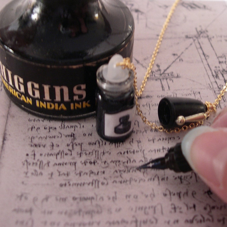 Op de afbeelding: Een ketting met een miniatuur zwart inktflesje als hanger aan een gouden ketting. De hanger is een gedetailleerde replica van een inktflesje met een wit etiket. Een grotere Higgins American India Ink fles staat op de achtergrond.