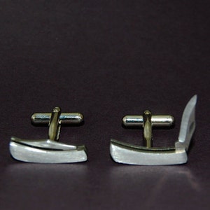 May include: A pair of silver-toned cufflinks, one closed and one open. The cufflinks have a rectangular design with a curved edge and a silver bar connecting the two sides. The background is a dark brown color.
