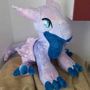 Könnte beinhalten: Ein Plüschdrache in Lila und Blau mit einem hellblauen Bauch und blauen Krallen. Der Drache hat einen weißen Fleck auf der Nase und ein großes, leuchtend blaues Auge.