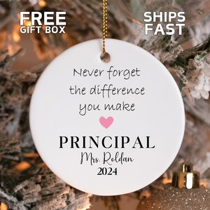 May include: A white ceramic ornament with gold string for hanging. The ornament has the text "Never forget the difference you make" with a pink heart, the word "PRINCIPAL", the name "Mrs. Roldan", and the year "2024".