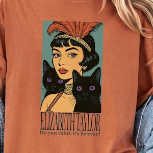 May include: Burnt orange t-shirt featuring a graphic of Elizabeth Taylor with two black cats. The image includes the text "ELIZABETH TAYLOR" and "Do you think it's forever?" The shirt has rolled-up sleeves and is paired with a denim jacket.