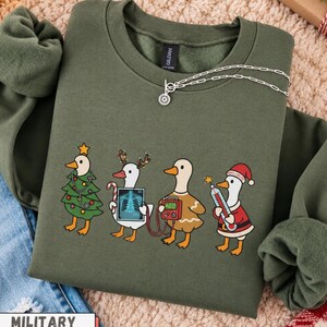 May include: A military green crewneck sweatshirt featuring a graphic of four ducks dressed in medical-themed holiday attire. One duck is a Christmas tree with a stethoscope, another holds an X-ray, one wears a sweater with an AED, and one is Santa.