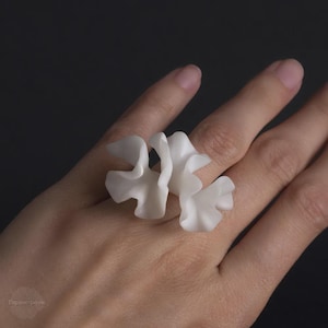 Könnte beinhalten: Ein weißer, blütenförmiger Ring mit einem zarten, gerüschten Design. Der Ring besteht aus einem glatten, undurchsichtigen Material und wird an einem Finger getragen. Der Hintergrund ist dunkelgrau.