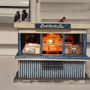 Puede incluir: Un modelo en miniatura de un quiosco de prensa con una fachada a rayas azules y blancas. El letrero sobre la ventana dice "Союзпечати". Las ventanas est&aacute;n llenas de revistas y peri&oacute;dicos en miniatura. Hay tres p&aacute;jaros peque&ntilde;os posados en el techo.