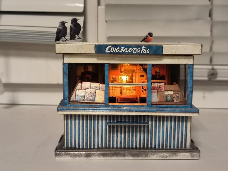 Puede incluir: Un modelo en miniatura de un quiosco de prensa con una fachada a rayas azules y blancas. El letrero sobre la ventana dice "Союзпечати". Las ventanas est&aacute;n llenas de revistas y peri&oacute;dicos en miniatura. Hay tres p&aacute;jaros peque&ntilde;os posados en el techo.