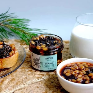 Puede incluir: Un tarro de mermelada de piñas con piñones, un cuenco pequeño de mermelada, un vaso de leche y un pastel en un plato. El tarro tiene una etiqueta verde con texto y una ilustración de piña.