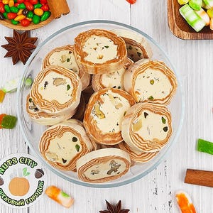 Puede incluir: Vista aérea de un cuenco de cristal lleno de pasteles en rodajas y en capas con un relleno cremoso y nueces. Caramelos, especias y una pegatina con texto están esparcidos alrededor del cuenco sobre una superficie de madera blanca. Los pasteles son de color marrón claro y blanco.