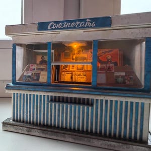 Puede incluir: Un modelo en miniatura de un quiosco de prensa sovi&eacute;tico con una fachada a rayas azules y blancas. El letrero sobre la ventana dice "Союзпечати" en escritura cir&iacute;lica. El interior del quiosco es visible, con estantes llenos de revistas y peri&oacute;dicos.