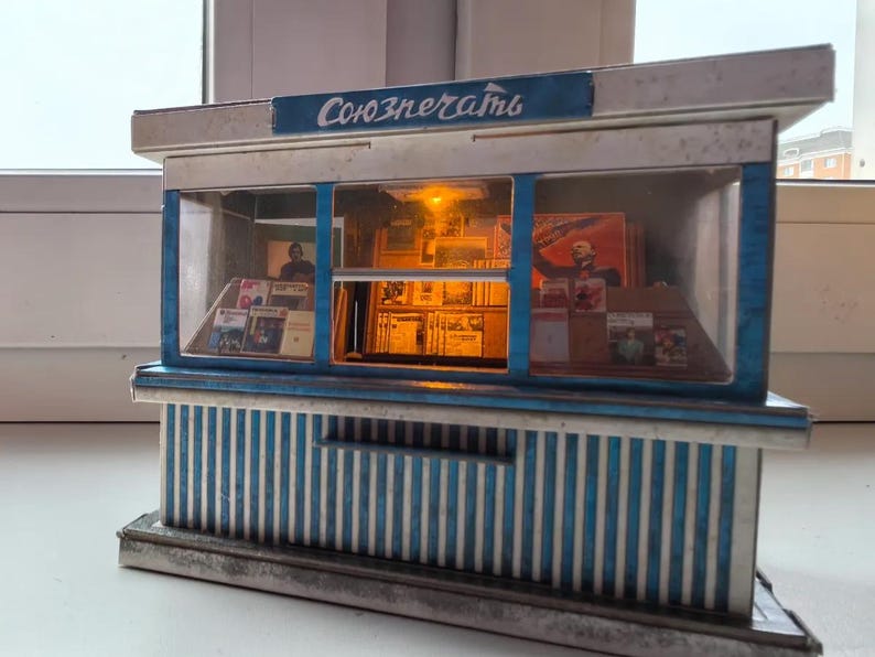 Puede incluir: Un modelo en miniatura de un quiosco de prensa sovi&eacute;tico con una fachada a rayas azules y blancas. El letrero sobre la ventana dice "Союзпечати" en escritura cir&iacute;lica. El interior del quiosco es visible, con estantes llenos de revistas y peri&oacute;dicos.
