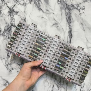 Puede incluir: Maqueta en miniatura de un edificio de apartamentos de varios pisos con numerosas ventanas y balcones. La fachada del edificio es principalmente gris con acentos coloridos en los balcones. El modelo está sostenido por una mano sobre un fondo con vetas de mármol.