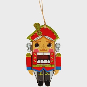 Puede incluir: Adorno navideño de madera con forma de cascanueces. Presenta un abrigo rojo, un sombrero verde y una cara detallada con la boca abierta. Se adjunta una cuerda para colgarlo. El diseño es colorido y festivo.