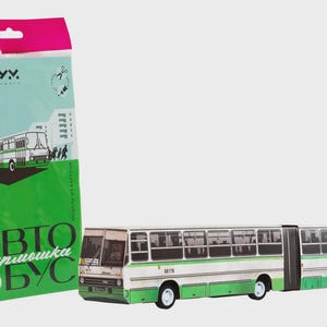 May include: A white and green articulated bus model with a matching package. The bus has multiple windows and a detailed design. The package has a pink top and green bottom with text and a bus illustration.
