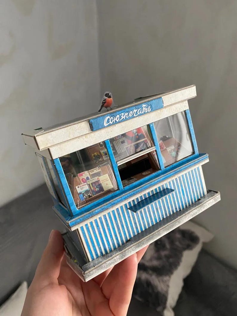 Puede incluir: Un modelo en miniatura de un quiosco azul y blanco a rayas con un letrero que dice "Союзпечати" y un peque&ntilde;o p&aacute;jaro posado en la parte superior.