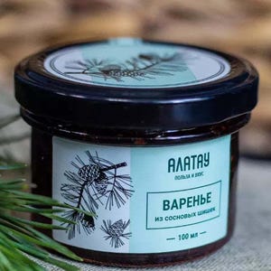 Puede incluir: Un tarro de mermelada de piñas con tapa negra y etiqueta con ilustraciones de piñas y texto. La etiqueta dice "Alatay" y "Mermelada de piñas", con un volumen de 100 ml.