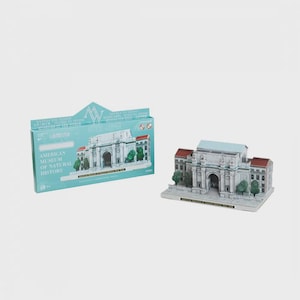 May include: A miniature model of the American Museum of Natural History in New York. The model is white with a blue roof and red accents. The model is next to its packaging, which features the museum's name and logo.