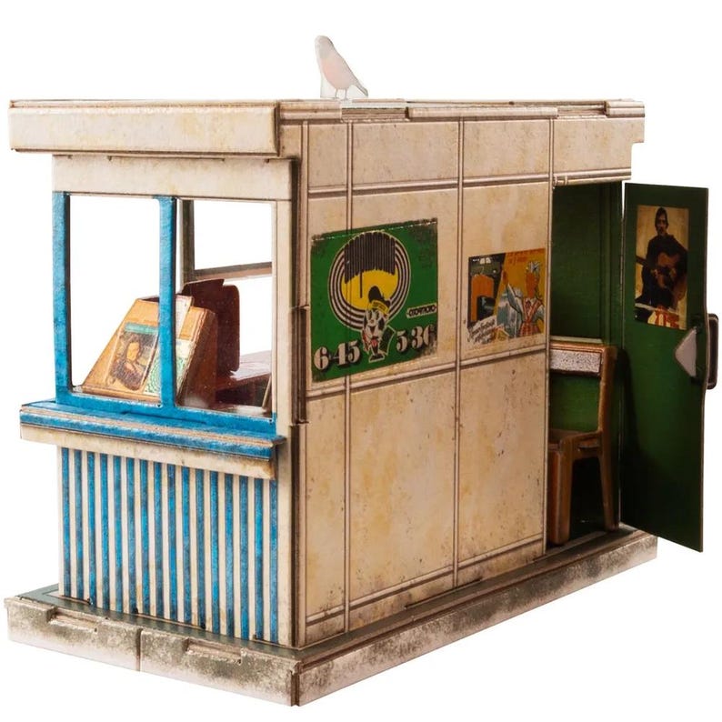 Puede incluir: Un modelo de cart&oacute;n en miniatura de una peque&ntilde;a tienda con una fachada a rayas azules y blancas, una puerta verde y un letrero que dice "645 536".
