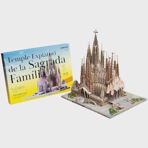 Op de afbeelding: Een gedetailleerd architectonisch model van de Sagrada Família-kathedraal, met een doos met de naam van het gebouw in het Spaans. Het model is beige en grijs, met ingewikkelde details. De doos heeft een blauwe en gele achtergrond.