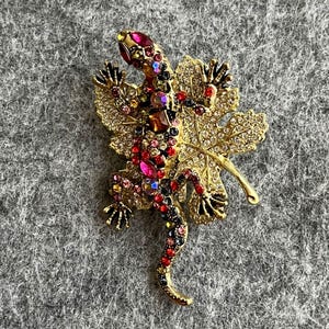 Può includere: Spilla dorata a forma di lucertola, impreziosita da cristalli rossi, rosa e trasparenti. La lucertola poggia su una foglia dorata, creando un accessorio ispirato alla natura.