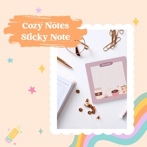 May include: A pink sticky note pad with a grid pattern and a cute illustration of coffee cups. The text "Cozy Notes" is printed on the top of the pad.