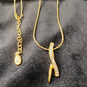 May include: A gold-toned necklace with a pendant featuring a curved design adorned with clear crystals. The necklace has a delicate chain and an adjustable clasp. The pendant is a striking accent piece.