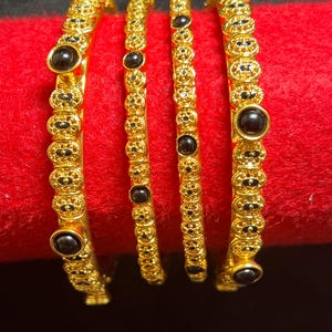 May include: Four gold bangles with detailed patterns and black gemstone embellishments. The bangles are presented on a red surface, highlighting their decorative design and polished appearance. The jewellery is suitable for special occasions.